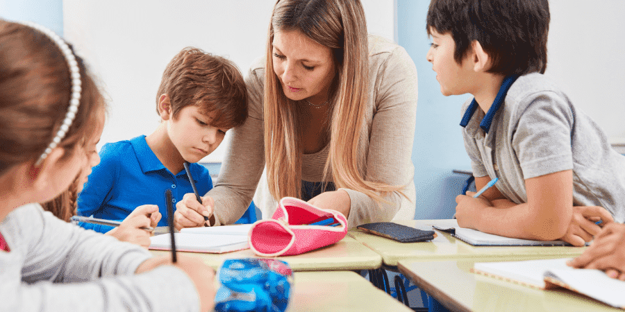 Eine Lehrerin beugt sich über einen Schreibtisch und hilft einem Jungen in einem blauen Shirt bei seinen Aufgaben, während andere Kinder zusehen.